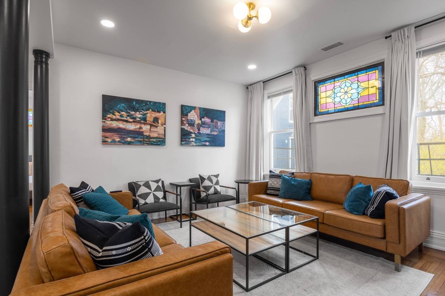 Downstairs living room — tan leather sofas, a gallery wall, and a stained glass window.
